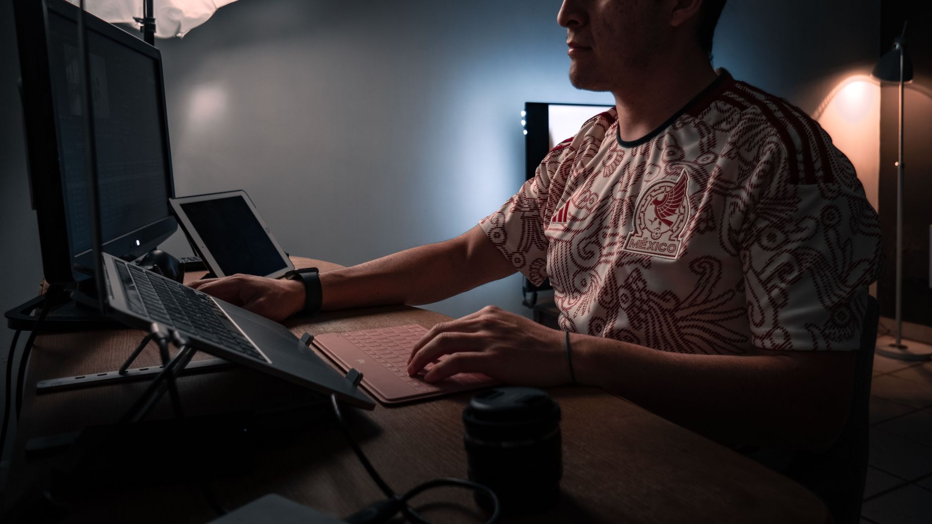 Mężczyzna w wzorowanej koszulce pracujący na laptopie i tablecie w słabo oświetlonym pomieszczeniu