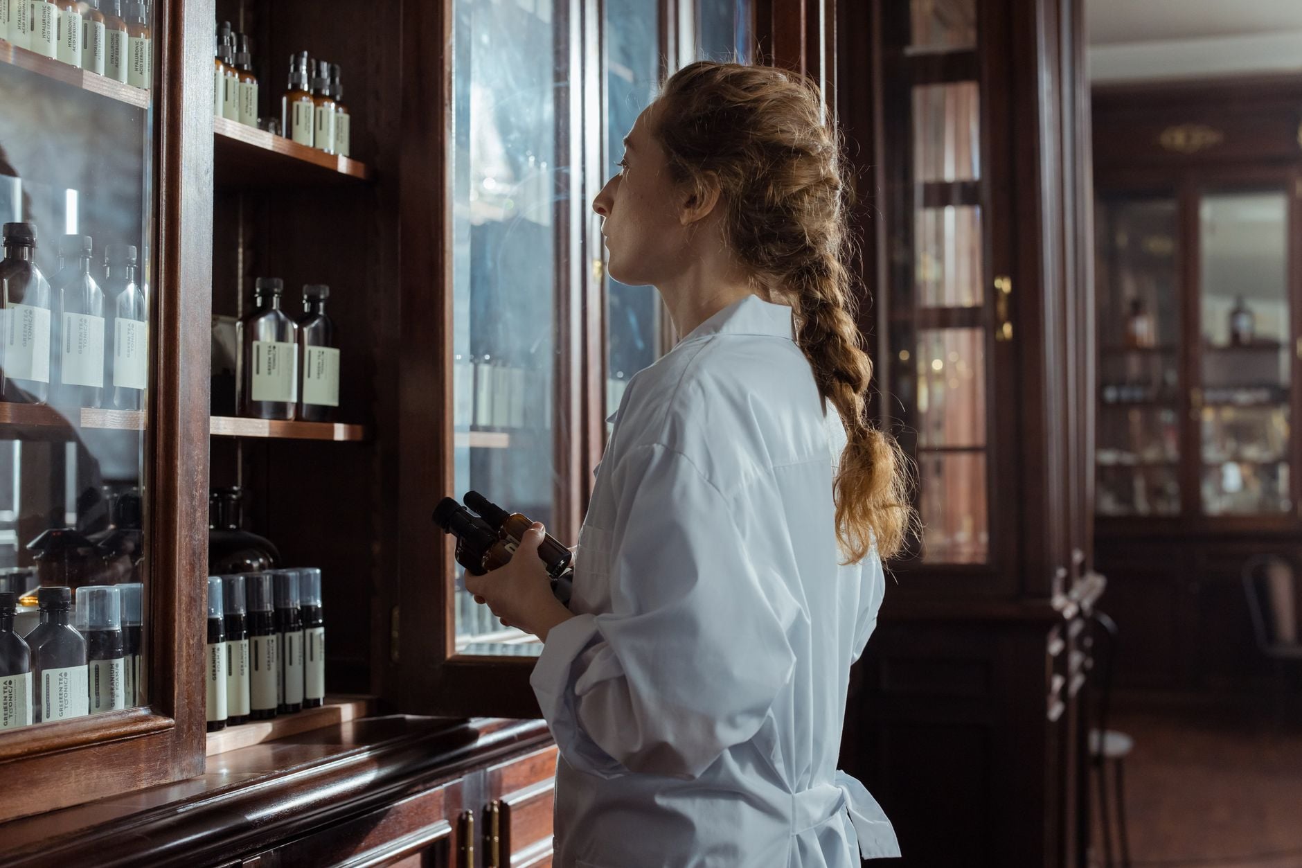 Kobieta w białym kitlu przygląda się butelkom produktów na drewnianych półkach z przeszklonymi drzwiami.